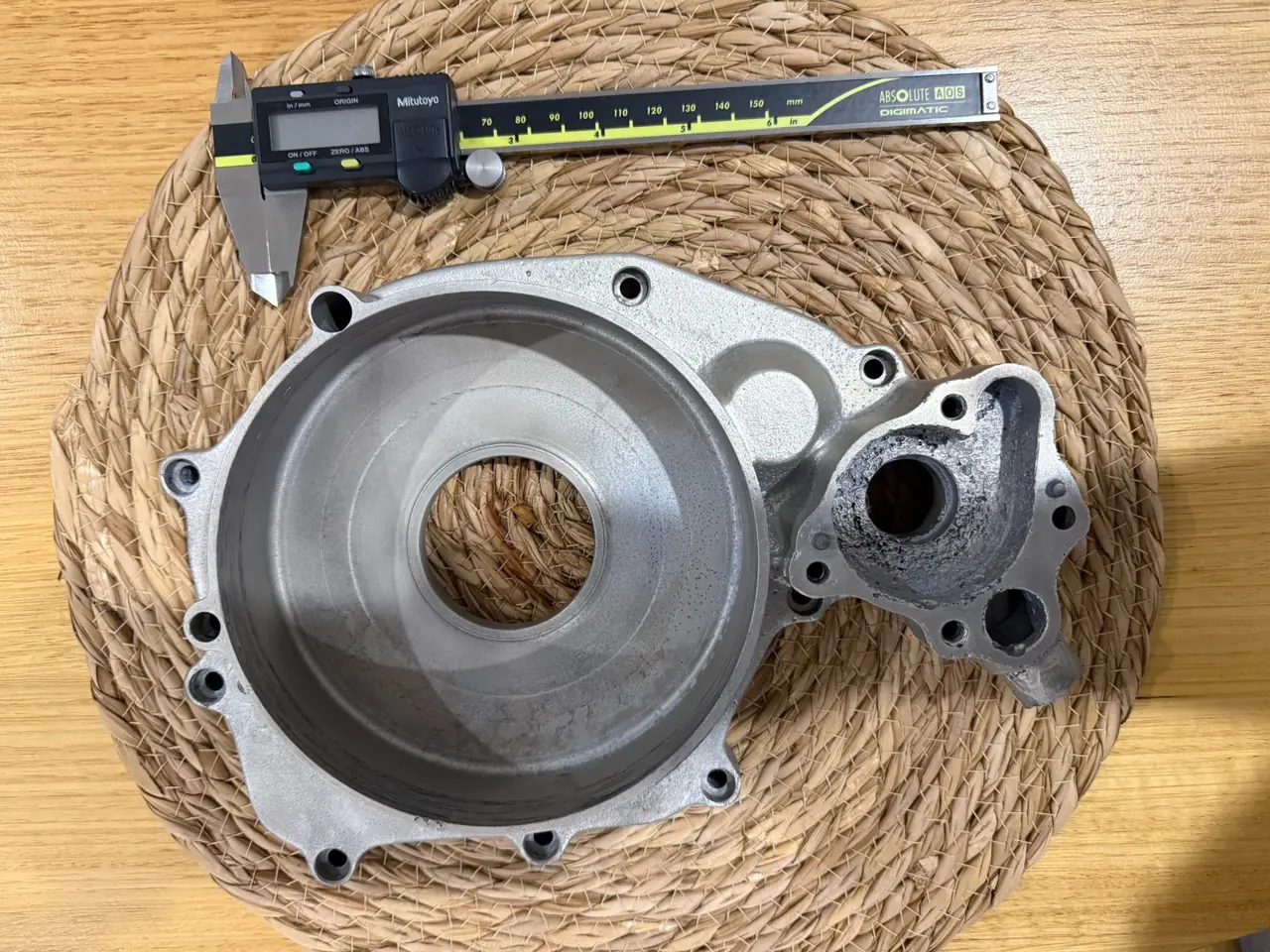 A 125cc motorcycle clutch cover photographed face-up on a table, a smaller cast component resting on it and a digital caliper laid alongside for scale — the aluminium casting EKINSUN was asked to reproduce in 6061