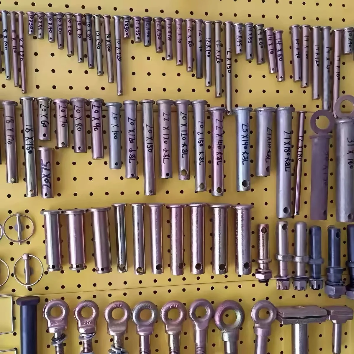 Workshop sample board of yellow zinc plated clevis and lock pins hung in rows on a pegboard, each pin marked in ink with its diameter and length from 8 x 100 up to 30 x 120 mm, with lynch pins and forged eye bolts along the bottom