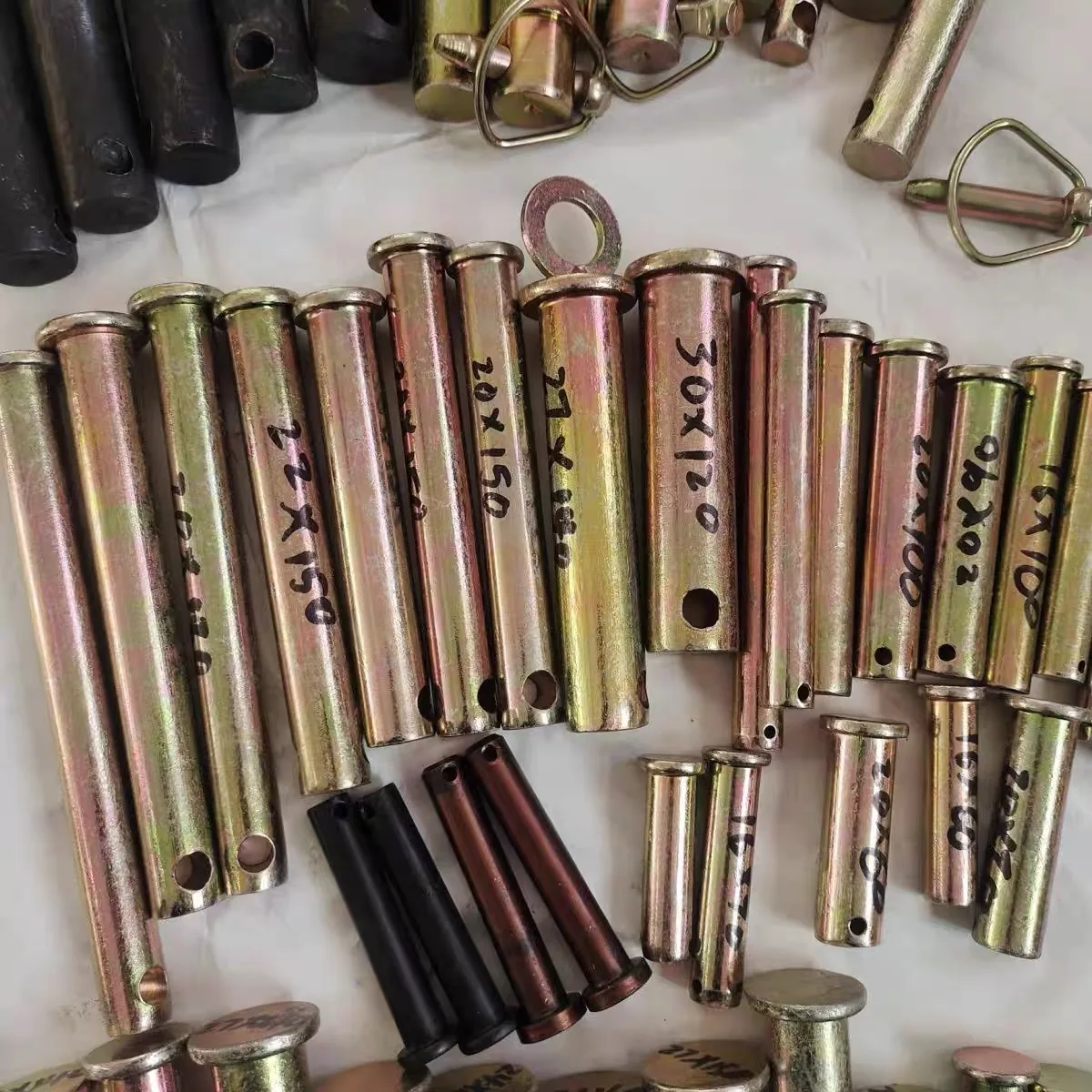 Loose group of machined lock pins in several finishes on a white cloth, yellow zinc plated pins marked 20 x 150, 22 x 150, 27 x 180 and 30 x 120 alongside black oxide pins, a ring-head pin and a wire lynch pin