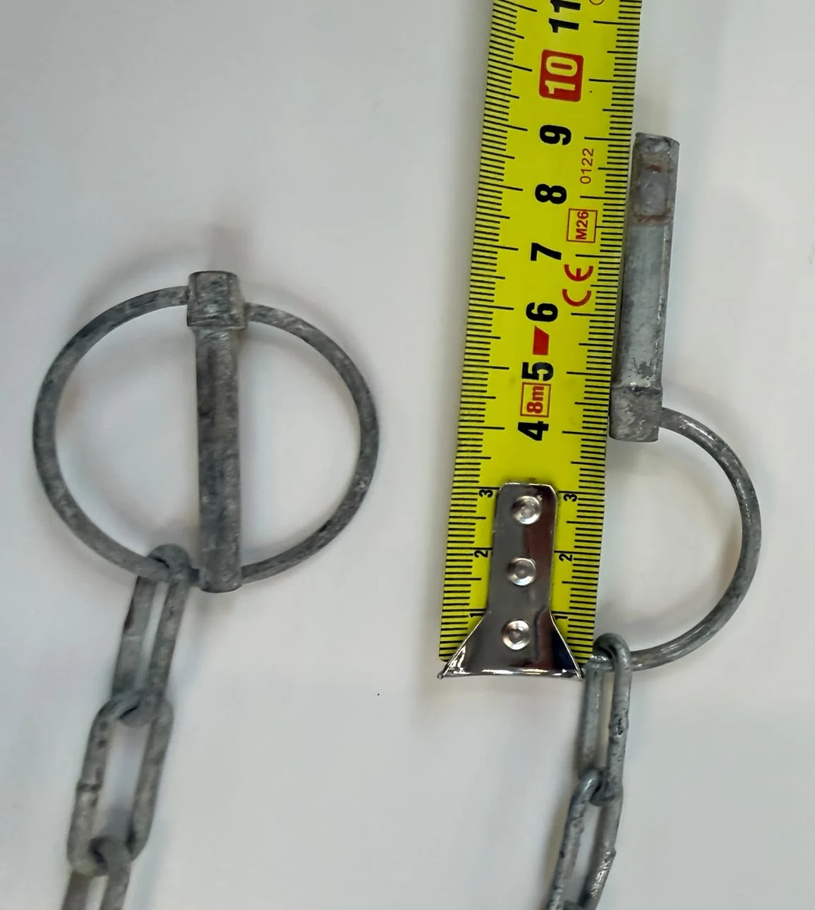 Two hot-dip galvanised lynch pins, each with a retaining chain hanging from its hinged ring, laid on a white surface with a folding rule set alongside one of them to read its length