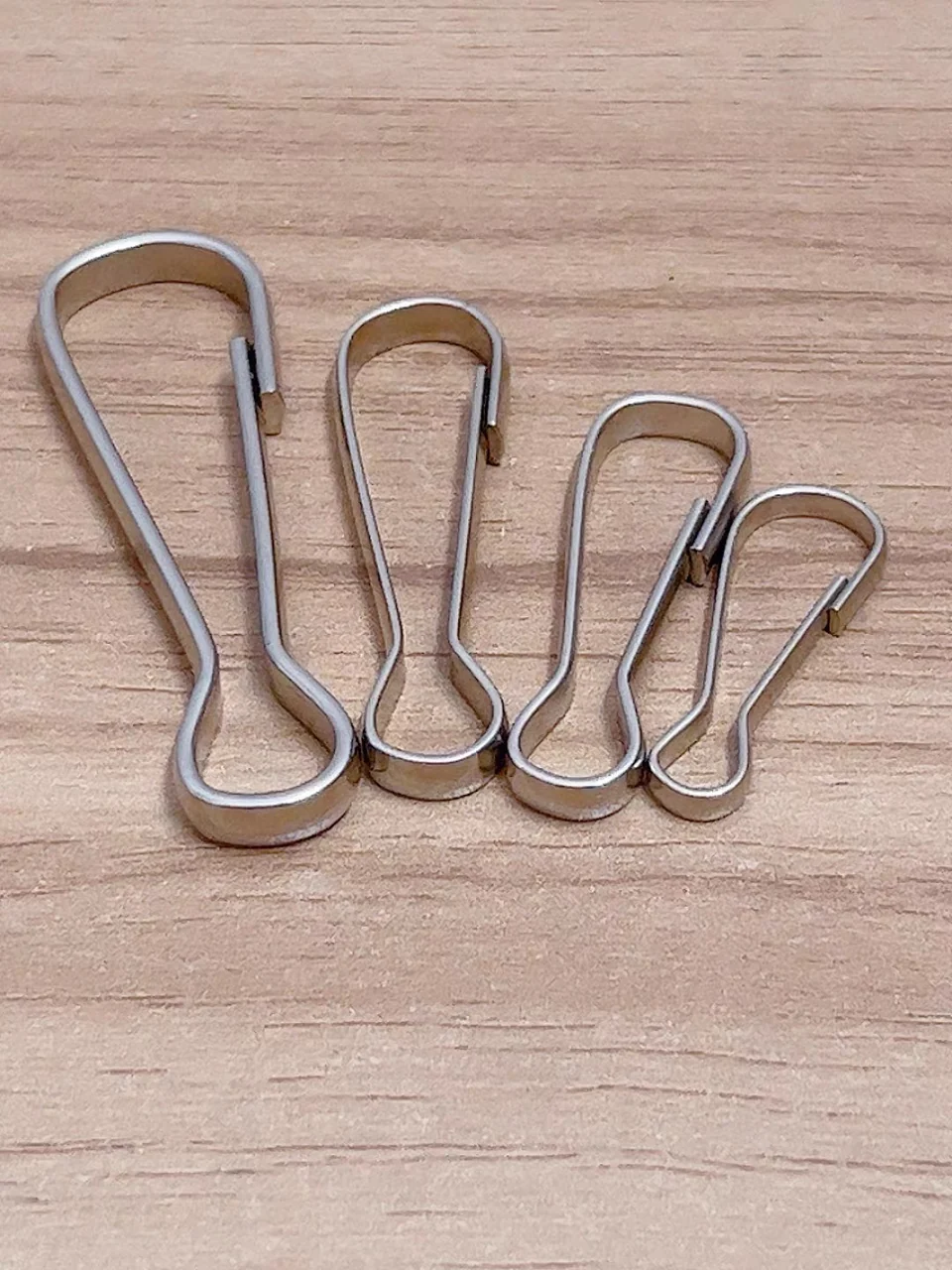 Four stainless steel spring snap hooks of decreasing size laid on a wooden bench, each a bent wire loop with a sprung gate at the narrow end and an eye at the wide end