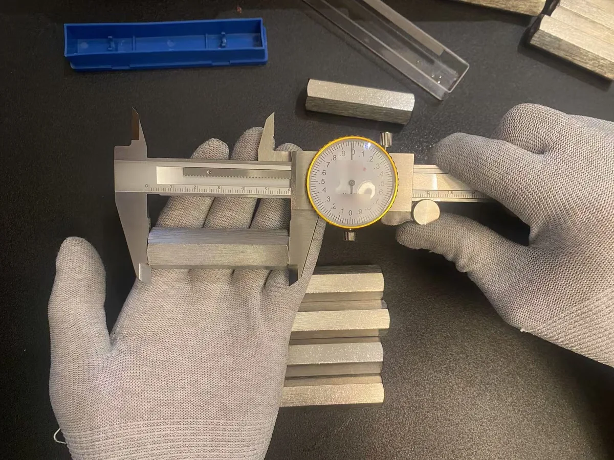 Dial caliper measuring the across-flats of a 304 stainless hex coupling nut, a row of finished nuts beside it