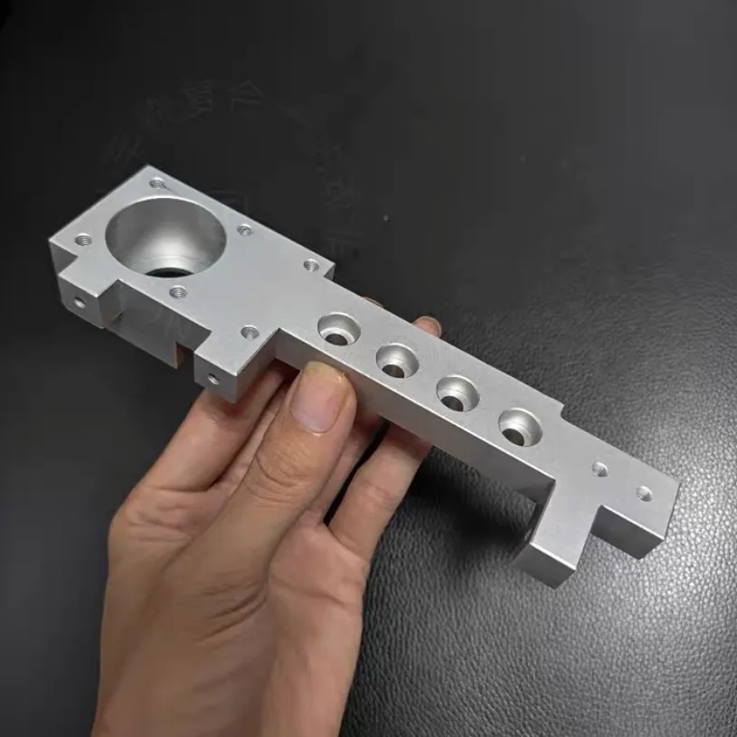 Hand holding a long aluminium bracket with a stepped bore at one end and a row of counterbored holes