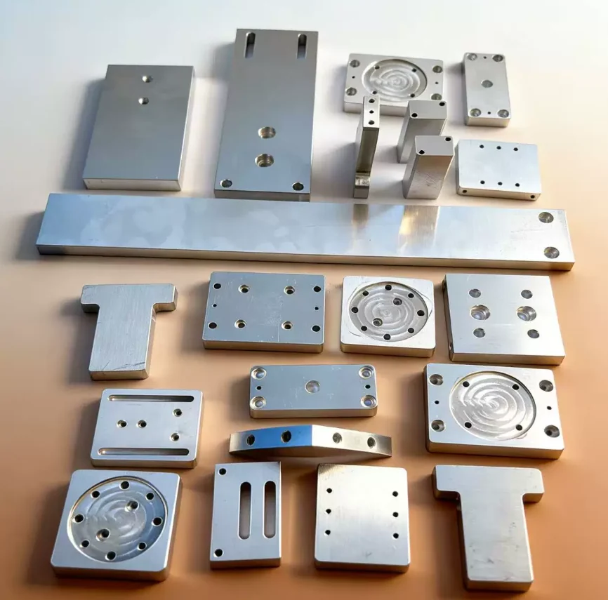 Machined aluminium blocks, flat plates with bolt patterns, T-shaped brackets, a long rail and circular plates with spiral grooves, laid out together