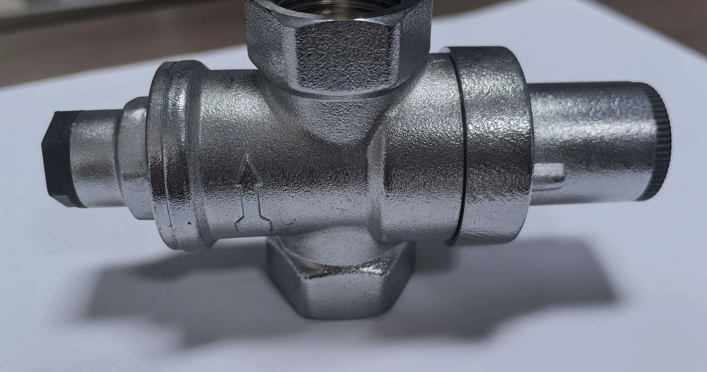A DN15 piston-type pressure reducing valve in nickel-plated brass lying on white paper: G1/2 female ports on both sides, the flow arrow cast into the body, the knurled black adjusting cap on the right and the black bottom plug on the left