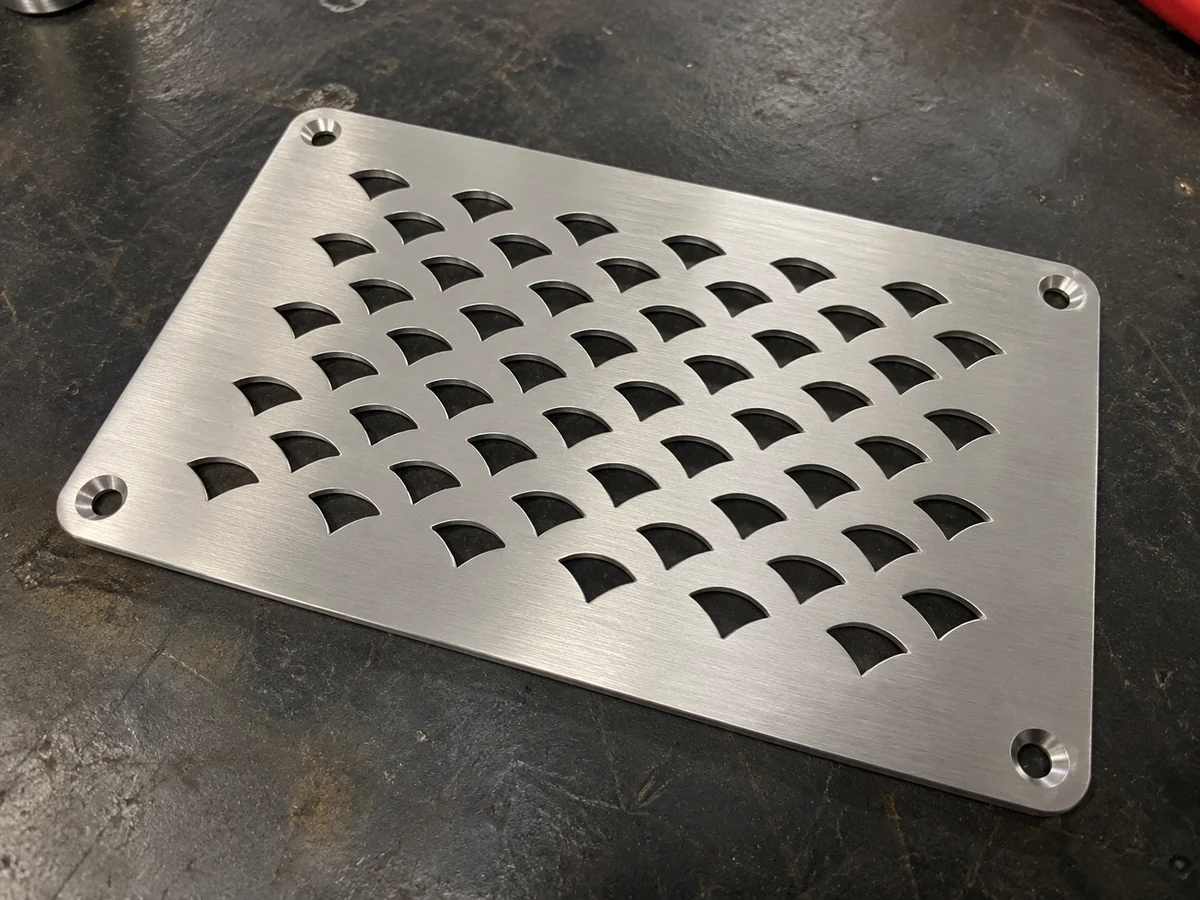 Brushed stainless steel plate with rows of scale-shaped cutouts in a staggered pattern and four countersunk corner holes, on a workshop floor