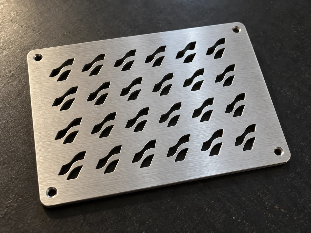 Brushed stainless steel plate with rows of wave-shaped cutouts and four chamfered corner holes, lying on a dark workbench