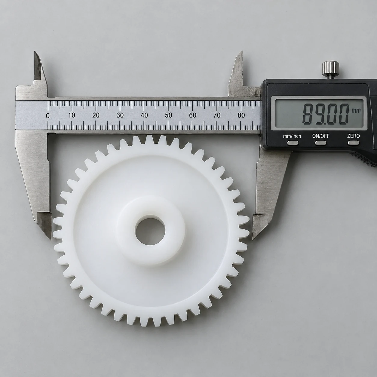 A digital caliper laid across a white nylon spur gear on a grey background, its jaws touching opposite tooth tips and the display reading 89.00 mm, with the gear's teeth, hub and bore all in frame and countable