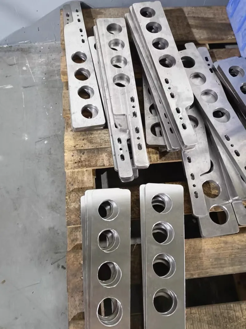 Stacks of machined metal plates with rows of large chamfered holes and small slots on a wooden pallet