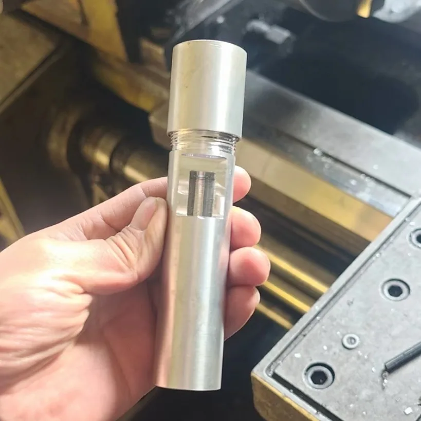 Hand holding a machined tube housing with a threaded cap and a clear section showing the part inside, in front of a machine table
