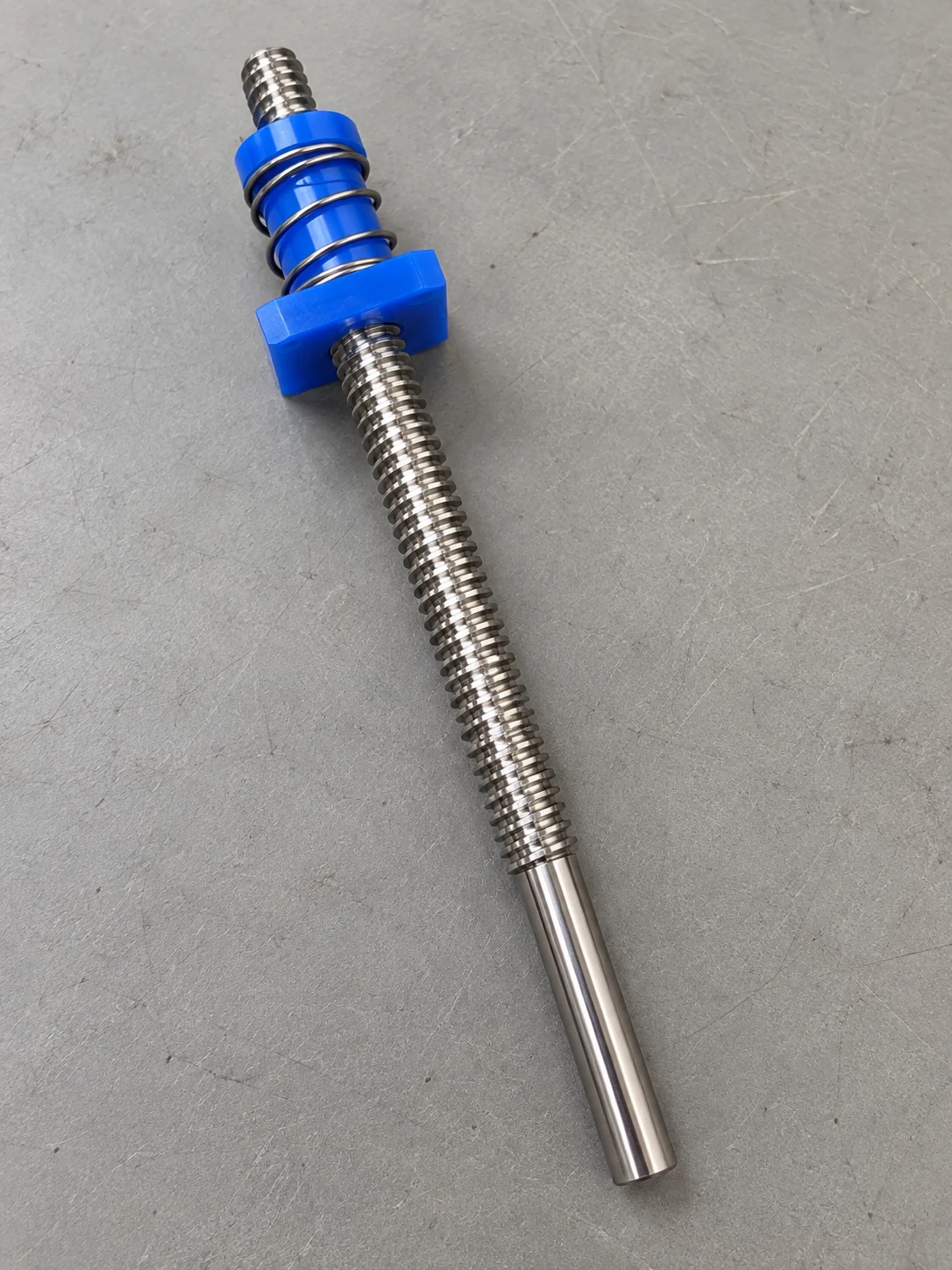 Stainless trapezoidal lead screw with a blue plastic anti-backlash nut, two nut halves held apart by a coil spring, plain end below the thread