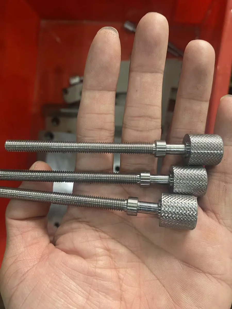 Trois vis moletées longues usinées sur mesure : tête à moletage croisé, longue tige filetée et collet lisse, présentées en main à l'atelier — EKINSUN
