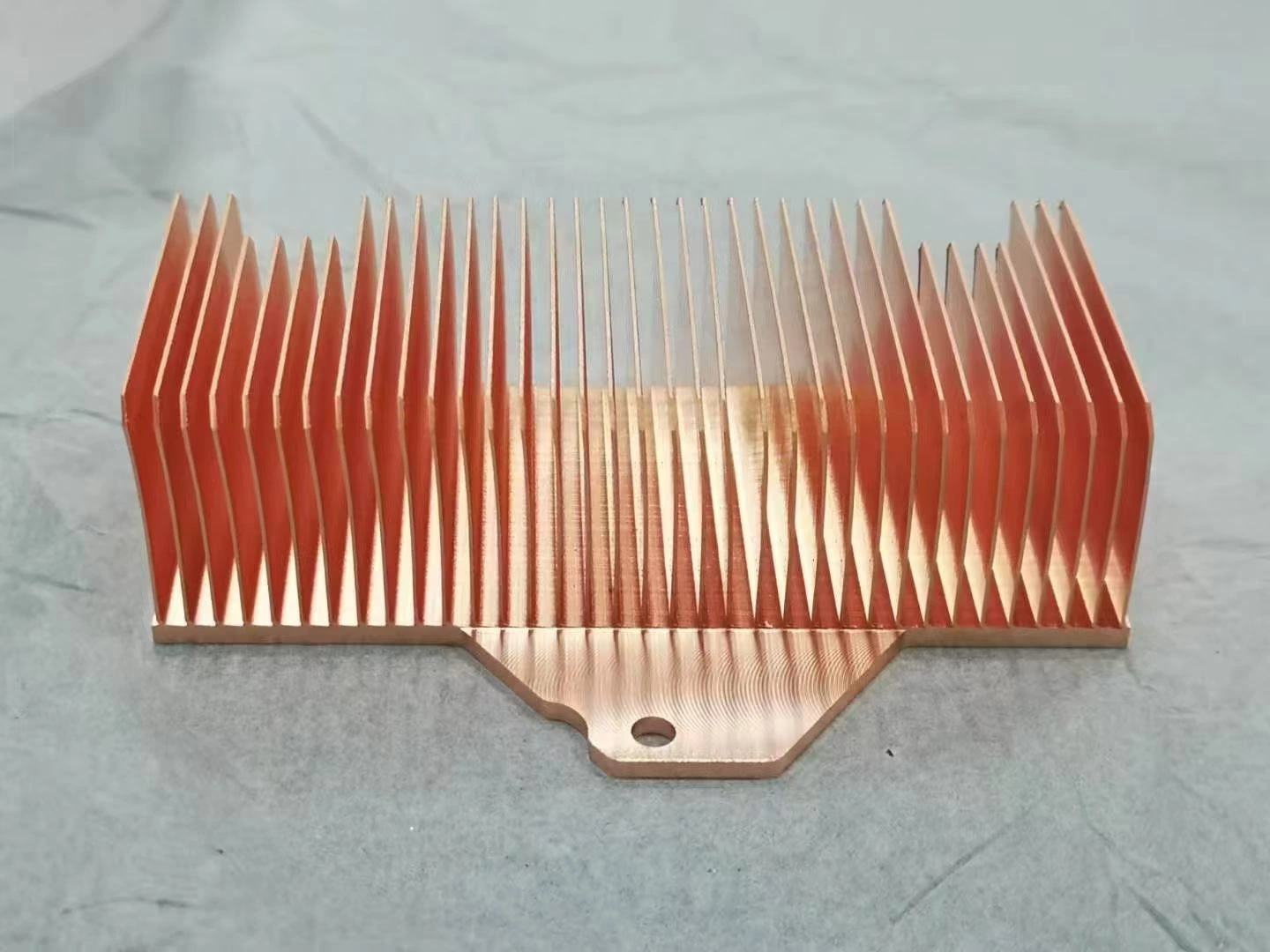 Side view of a copper skived fin heat sink, tall thin fins tapering towards the tip standing on a base profiled with chamfered corners and a mounting hole