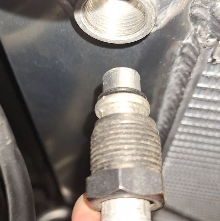 Customer photograph of an oil cooler line fitting held below the female threaded port of a radiator tank: the fitting shows a smooth nose with an O-ring, a wider collar, a run of external thread and a hex, with the oil line entering from below
